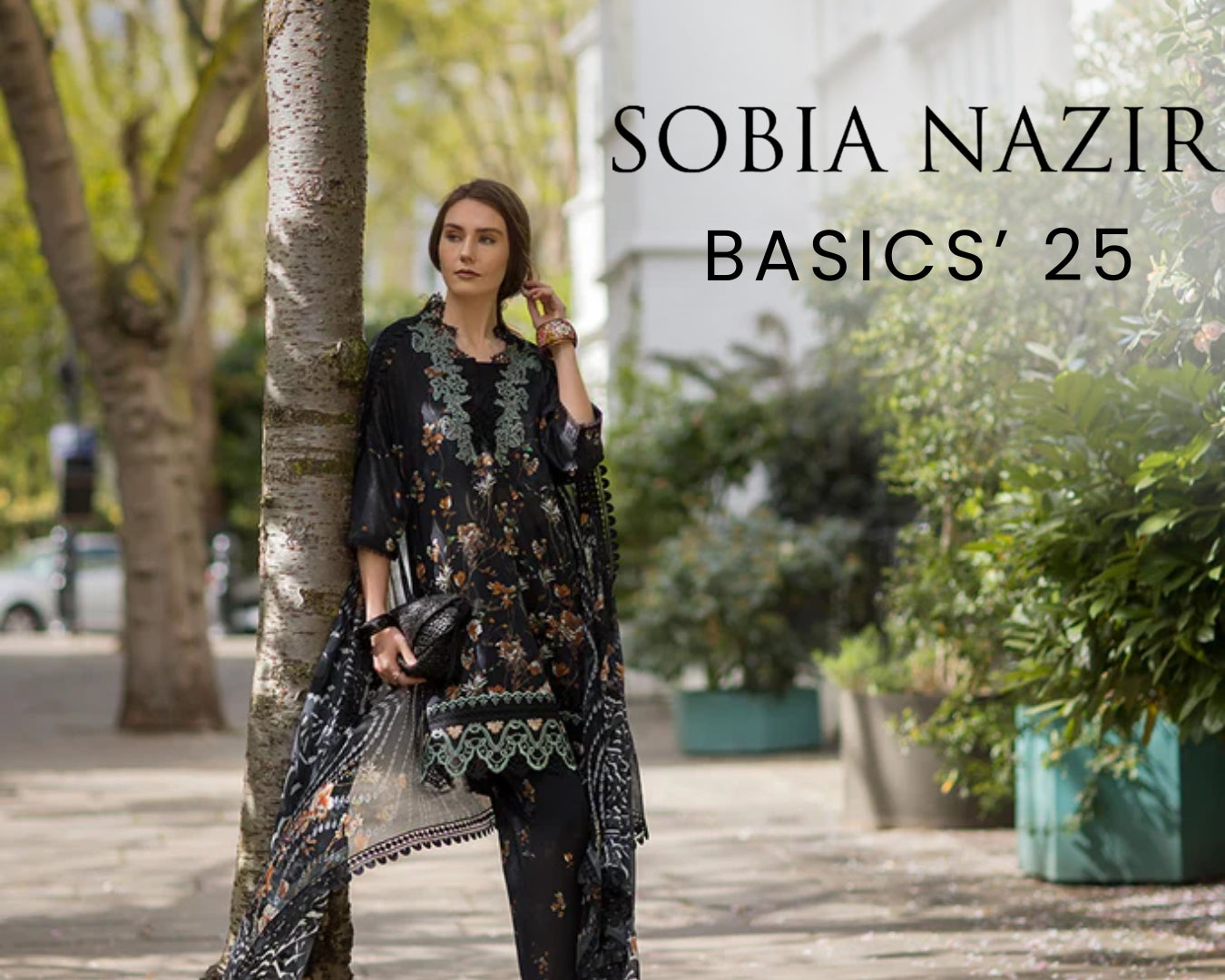 Woman in a black floral outfit standing next to a tree with 'Sobia Nazir Basics' 25 text.